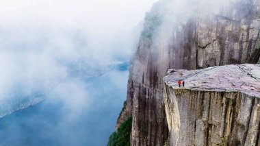 Maceracılar, aşağıdaki nefes kesen fiyortlara tepeden bakan görkemli Preikestolen Cliff 'in üzerinde dururlar. Sis dramatik manzarayı silip süpürüyor büyüleyici bir atmosfer yaratıyor..