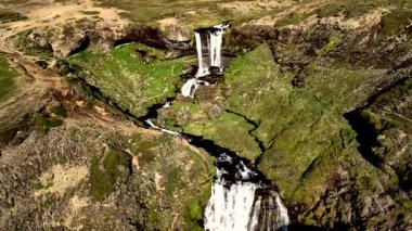 İzlanda 'daki Grundarfoss şelalesinin nefes kesici güzelliğini tecrübe edin. Suyun zarif bir şekilde uçurumlardan aktığı, canlı yeşillikler ve engebeli araziyle çevrili parlak mavi bir gökyüzü altında..