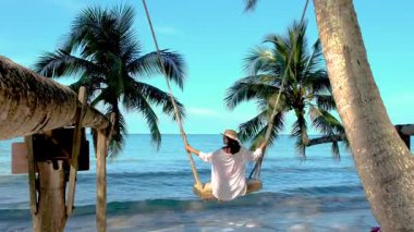 Bir kadın, etrafında palmiye ağaçları ve Tayland 'daki Koh Kood Adası' nın sakin okyanus dalgalarıyla çevrili bir sahilde salıncakta sallanmaktan zevk alır. Tropik cennet atmosferlerini sergilemek için mükemmel..