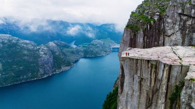 Ziyaretçiler, Norveç 'in dramatik fiyort manzarasına hayret ederek Preikestolen Cliff' in kenarında duruyorlar. Sakin mavi sular engebeli yeşil tepelerle tezat oluşturuyor. Bulutlu bir gökyüzü atmosfere ekleniyor.