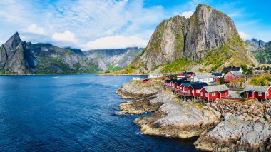 Aydınlık bir günde Hamnoy 'un kayalık kıyı şeridi renkli kulübelerle doludur. Arka planda görkemli dağlar yükselir, sakin sularla çarpıcı bir kontrast yaratır..