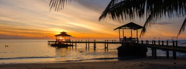 Güneş Koh Kood Adası 'ndaki sakin bir rıhtımın arkasında batıyor. Gökyüzüne canlı renkler saçıyor ve sakin sulara yansıyor. Tayland 'da mükemmel bir kaçış için kıyıda hafif dalgalar var.