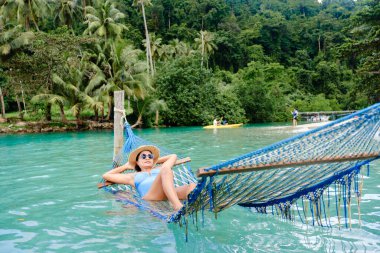 Bir kadın, Tayland 'ın Koh Kood kentindeki verimli tropik yeşilliklerle çevrili berrak turkuaz suların üzerindeki bir hamakta uzanarak huzurlu bir öğleden sonranın tadını çıkarıyor. Kayakçılar yakınlarda süzülüyor, dingin sahneyi büyütüyorlar..