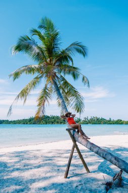 Tayland, Koh Kood 'da çarpıcı bir sahile uzanan bir ağaç gövdesine yaslanan bir gezgin. Berrak mavi sular ve canlı palmiye ağaçları mükemmel bir tropikal kaçış sahnesi yaratır..
