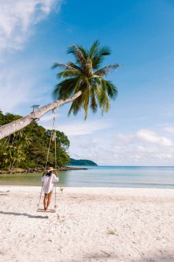 Koh Kood Adası 'ndaki sakin bir sahil sahnesinde uzun bir palmiye ağacının altında sallanan yalnız bir figür. Açık mavi gökyüzü ve durgun turkuaz sular dinlenmek için mükemmel bir atmosfer yaratır..