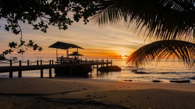 Güneş Koh Kood Adası 'nın üzerinde batarken gökyüzünü altın renkleri boyuyor. Palmiye yapraklarıyla çerçevelenmiş, sakin denize doğru uzanan büyüleyici bir ahşap iskele dinlenmek için mükemmel, huzurlu bir tropik cennet yaratıyor..