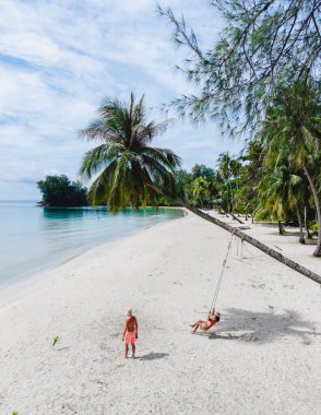 Bir çift beyaz erkek ve Asyalı kadın Koh Kood, Tayland 'da sakin bir sahilde mutlu mesut sallanırken bir diğeri sallanan palmiye ağaçlarının altında yürüyor..