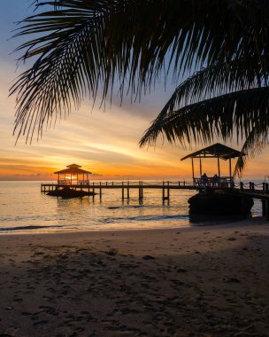 Tayland 'daki Koh Kood Adası' nda çarpıcı bir günbatımının tadını çıkar. Huzurlu sahil sahnesinde, sallanan palmiyeler ve canlı gökyüzü renkleriyle çevrili ahşap bir iskele ve huzurlu köşkler yer alıyor..