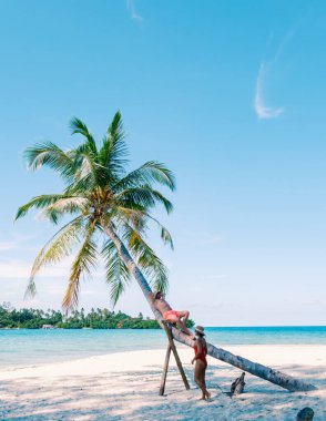 Çeşitli bir çift, temiz mavi bir gökyüzünün altında el değmemiş beyaz kumsallardaki bir palmiye ağacına tırmanarak Koh Kood Adası 'nın güzelliğini deneyimliyor. Sakin sular rahatlamaya ve maceraya davetiye çıkarır..