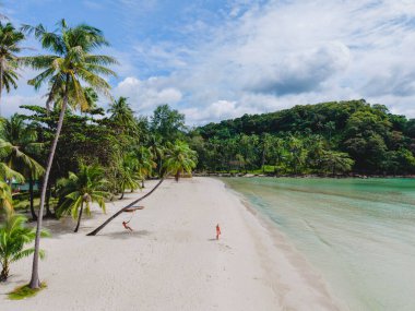 Tayland 'daki Koh Kood Adası' nın güzelliğini keşfedin, yumuşak beyaz kumların kristal berrak sularla buluştuğu yer. Bir çift adam ve bir kadın sahildeki palmiye ağacının altında salıncakta sallanıyorlar.