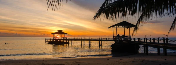 Güneş Koh Kood Adası 'ndaki sakin bir rıhtımın arkasında batıyor. Gökyüzüne canlı renkler saçıyor ve sakin sulara yansıyor. Tayland 'da mükemmel bir kaçış için kıyıda hafif dalgalar var.