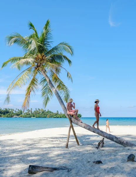 Beyaz bir adam, başka bir dişinin, sallanan palmiye ağaçları ve canlı mavi bir denizin yanında güneşin altında güneşlenmesine dikkat ediyor. Genç bir adam Tayland sahilinde başka bir kadını kontrol ediyor, Koh Kood.