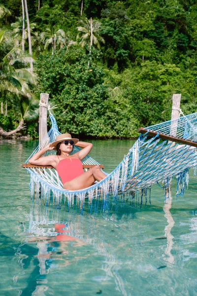 Bir kadın turkuaz suların üzerindeki direklerin arasına sarılı renkli bir hamakta uzanır, yemyeşil sularla çevrili. Sakin atmosfer, Koh Kood Tayland 'daki sıcak güneşin altında rahatlamaya davet ediyor.
