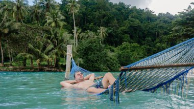 Tayland 'daki Koh Kood Adası' nın berrak turkuaz suları üzerinde asılı bir hamakta uzanan bir adam, arka planda yoğun yeşil yapraklarla huzurlu bir anın tadını çıkarıyor..