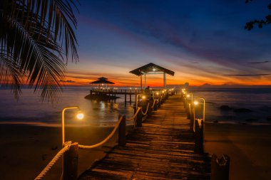 Alacakaranlık, Koh Kood Adası 'ndaki huzurlu iskelede güneş batarken gökyüzünü turuncu ve mor renklere boyuyor. Işıklar yolu aydınlatıyor, sakin suları yansıtıyor, sükuneti davet ediyor..