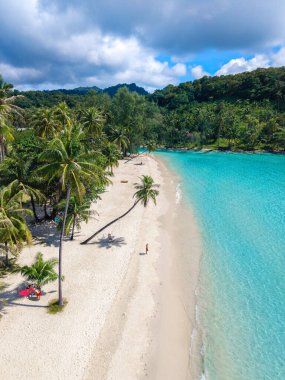 Beyaz kumlu plaj Koh Kood Adası 'nın turkuaz suları boyunca uzanır ve uzun palmiye ağaçları tarafından çerçevelenir. Güneş banyosu yapanlar açık mavi gökyüzünün altındaki renkli havlularda rahatlarlar, cennetin huzurlu güzelliğini yakalarlar..