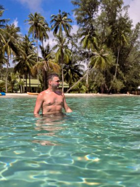 Bir adam Koh Kood Adası 'nın turkuaz sularında durmaktan zevk alır. Bereketli palmiye ağaçları, parlak mavi gökyüzünün altında yavaşça sallanarak tropikal cennet atmosferini güçlendirir..