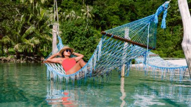Asyalı bir kadın, Tayland 'daki Koh Kood Adası' nın berrak suları üzerinde asılı duran renkli bir hamakta huzurlu anının tadını çıkarıyor. Bereketli yeşilliklerle çevrili, mükemmel bir tropikal kaçış.
