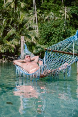 Tayland 'daki Koh Kood Adası' nın berrak sularında asılı duran renkli bir hamakta huzurlu bir anın tadını çıkaran bir adam. Etrafını saran yüksek hurma ağaçları, tropik mutluluğun vücut bulmuş hali..