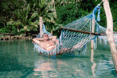 Bir adam, Koh Kood Adası 'nın dingin güzelliğinin tadını çıkarıyor. Berrak suların üzerindeki hamakta, yemyeşil tropikal bitki örtüsüyle çevrili. Tayland 'da dinlenmek ve gençleşmek için mükemmel bir yer.