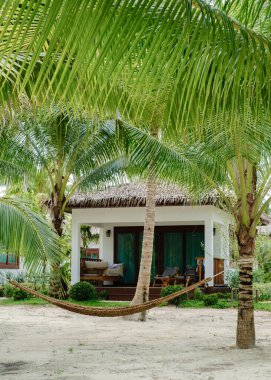 Koh Kood Adası 'ndaki büyüleyici bir villanın dışındaki palmiye ağaçları arasında sallanan sıcak bir hamak. Huzurlu ortam, ziyaretçileri yemyeşil ve kumlu Tayland kıyılarında dinlenmeye davet ediyor.