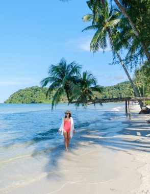 Tayland 'daki Koh Kood Adası' nın yumuşak kumlu sahillerinde, bereketli palmiye ağaçları ve nazik dalgalarla çevrili bir kadın zarif bir şekilde yürüyor. Açık mavi gökyüzü sakin atmosferi güçlendirir..