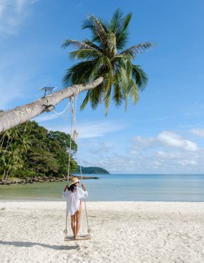 Koh Kood Adası 'ndaki güzel bir plaj sahnesinde uzun bir palmiye ağacının altında sallanan bir kadın görülüyor. Sakin mavi sular ve berrak gökyüzü Tayland 'da mükemmel bir tropik cennet atmosferi yaratır.