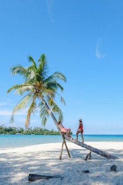 İki arkadaş Koh Kood Adası 'nın kumlu sahilinde bir palmiye ağacını inceliyor. Berrak gökyüzü ve turkuaz su bu tropik kaçış için mükemmel bir zemin oluşturur..