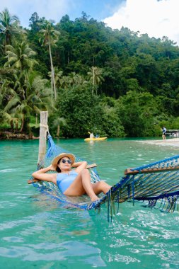 Renkli bir hamakta dinlenen bir kadın, Koh Kood Adası 'nın berrak turkuaz sularında hafifçe sallanıyor. Bereketli palmiye ağaçları ve sakin bir plaj bu güzel varış noktasının tropikal atmosferini arttırır..