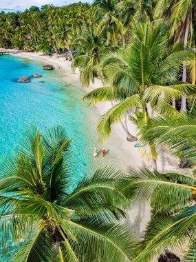 Kristal berrak sular Koh Kood Adası 'nın yumuşak kumlarını kaplarken palmiye ağaçları da mutlu bir sahil manzarası çiziyor. İki insan, Tayland 'daki sakin tropikal cennetin tadını çıkararak kıyı boyunca yürüyor.