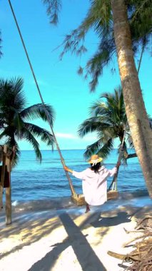 Koh Kood Adası 'ndaki palmiye ağaçlarının altında salıncaktan hoşlanan bir kadının huzurlu görüntüsü. Berrak mavi gökyüzü zümrüt sulara yansıyarak rahatlamak için mükemmel sakin bir atmosfer yaratır..