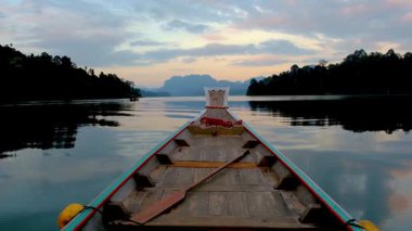 Geleneksel uzun kuyruklu tekne şafak vakti sakin sularda süzülürken Khao Sok Tayland 'ın huzurunu yakalayın. Göz kamaştırıcı yansımalar ve nefes kesici bir manzara sabah ışığında canlanıyor..