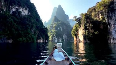Tayland 'daki Khao Sok Ulusal Parkı' nın huzurlu sularında süzül. Gün doğumunda uzun kuyruklu bir teknenin önünde elbise giymiş bir kadın.
