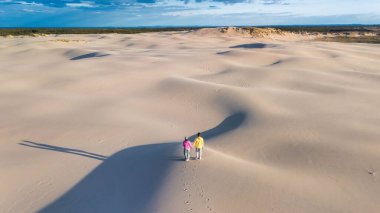 Danimarka 'nın geniş, dalgalı beyaz kum tepelerinde el ele dolaşan iki birey. Günbatımının sıcak altın ışığı uzun gölgeler bırakır Rabjerg Mile Dunes, Danimarka.