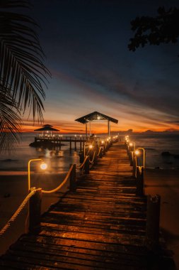 Tayland 'da Koh Kood Adası' nda güneş batarken sakin bir iskele yumuşak bir şekilde parlıyor. Sıcak turuncu ve mor renkler suyu yansıtıyor ve ziyaretçileri bu huzurlu akşam sahnesinin tadını çıkarmaya davet ediyor..