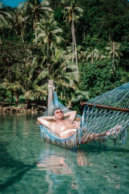 Koh Kood Adası 'nda bir adamın turkuaz suların üzerine asılmış bir hamakta sükunetin tadını çıkardığı sakin bir sahne. Güneş gibi parlayan palmiyeler yavaşça sallanarak tropik bir cennet yaratıyor..