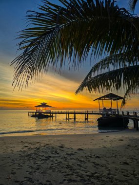 Güneş Koh Kood Adası, Tayland 'da batarken gökyüzünü altın renkleri kaplıyor. Berrak palmiye ağaçları tarafından çerçevelenmiş sakin sulara uzanan sakin bir iskele dinlenmek için huzurlu bir cennet yaratıyor..
