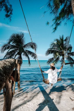 Koh Kood Adası 'ndaki sakin bir plaj sahnesinde palmiye ağaçlarından sallanan bir salıncak görülüyor. Bir kadın, Tayland 'ın çarpıcı turkuaz okyanus manzarasını izlerken hafif esintiden zevk alır.