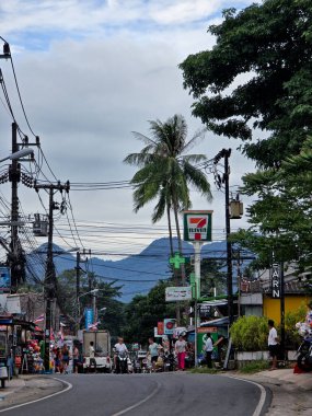 Koh Chang, Tayland, 30 Ekim 2025, hareketli bir sokak sahnesi, 711 dükkanının, insanların ve etrafındaki dağların arasında yerel hayatı gözler önüne seriyor..