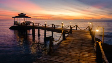 Güzel bir günbatımı, Koh Kood Adası 'nın sakin sularının üzerinde canlı renklerle gökyüzünü boyar. Ahşap iskele denize doğru uzanıyor, Tayland 'da dinlenmek için mükemmel huzurlu bir atmosfer yaratıyor.