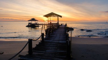 Güneş Koh Kood Adası Tayland 'ın sakin sularında batarken altın renkler gökyüzünü aydınlatıyor. Kırsal ahşap iskele ufka doğru uzanıyor, akşam yürüyüşlerini ve huzurlu yansımaları davet ediyor..