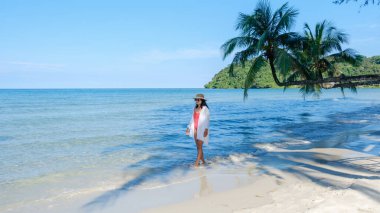 Bir kadın Tayland 'daki Koh Kood Adası' nın yumuşak kumlu plajında huzurlu bir yürüyüşün tadını çıkarıyor. Berrak mavi sular, bereketli yeşillikler ve parlak güneşin altında sallanan palmiyeler tarafından çerçevelenmiş sonsuz bir şekilde uzanır..
