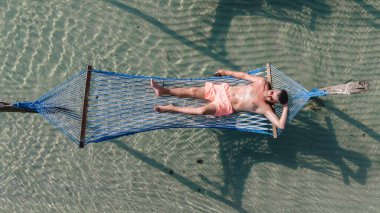 Tayland 'ın köpüklü suları üzerinde asılı duran hamakta dinlenen bir adamın sakin görüntüsü. Palmiye ağaçları yavaşça sallanırken güneş ışığı yüzeyde dans eder.