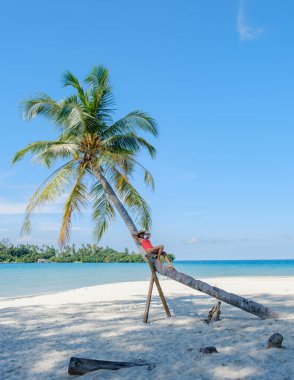 Koh Kood Adası 'nda sakin bir sahne, Asyalı bir kadının eğimli bir palmiye ağacında uzandığını gösteriyor. Açık mavi gökyüzü ve turkuaz sular Tayland 'da mükemmel bir tropikal kaçış yaratır.