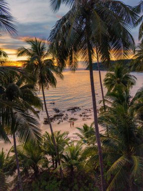 Gün batımında Koh Kood Adası 'nın sakin atmosferini hissedin. Altın renkler sakin suları yansıtıyor palmiye ağaçları usulca sallanırken kumsalda yürüyen bir çift kadın ve erkek