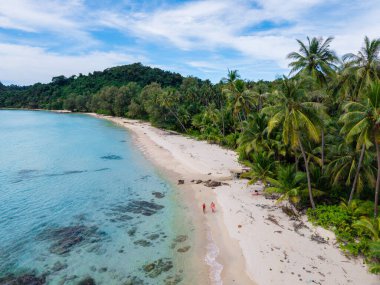 Canlı yeşil palmiye ağaçlarıyla çevrili yumuşak kumlu plajların sergilendiği Koh Kood Adası 'nın sakin bir manzarası. Tayland 'da kumsalda yürüyen bir çift erkek ve kadın, yukarıdan bir drone ile görülmüşler.