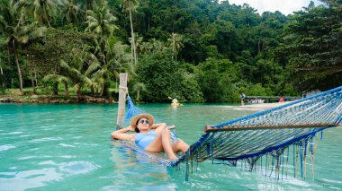 Bereketli yeşillikler ve Tayland 'daki Koh Kood Adası' nın turkuaz sularıyla çevrili bir hamakta uzanan bir kadının huzurlu tropikal atmosferinin tadını çıkardığı sakin bir manzara gözler önüne seriliyor..