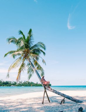 Adamın biri eğilmiş bir palmiye ağacına yaslanmış Tayland 'daki Koh Kood Adası' nın sıcak güneşinin tadını çıkarıyor. Kristal gibi berrak sular ve güzel bir sahil mükemmel bir tropikal kaçış yaratır..