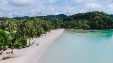 Tayland 'ın Koh Kood Adası' ndaki sakin plajda yumuşak kumlar, sallanan palmiye ağaçları ve turkuaz sular sergileniyor. Ziyaretçiler kıyı boyunca dinleniyor, açık bir gökyüzü altında tropik cennetin tadını çıkarıyorlar..