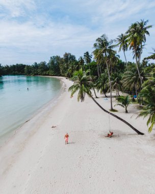 Tayland 'daki Koh Kood Adası' nın dingin güzelliğini görün. Huzurlu bir sahil yumuşak beyaz kum ve sallanan palmiye ağaçlarına sahiptir. Bir çift adam ve bir kadın sahildeki palmiye ağacının altında salıncakta sallanıyorlar.